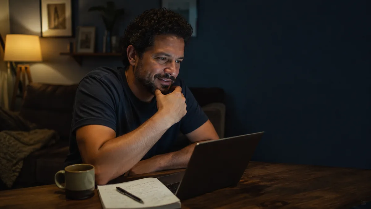 Homem sentado à mesa de casa à noite, estudando no notebook com um caderno de anotações ao lado
